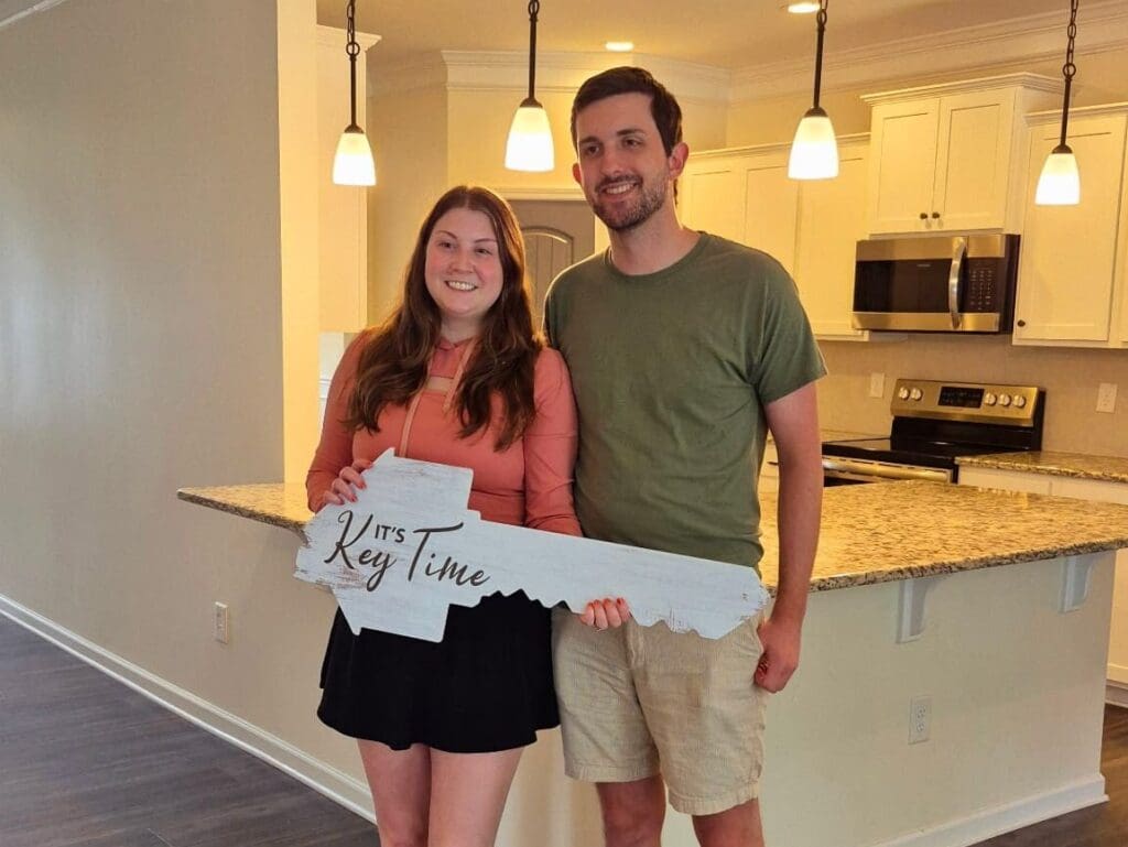 A smiling couple stands in a new home's kitchen, holding a large decorative key with "It's Key Time" written on it, conveying excitement and happiness.